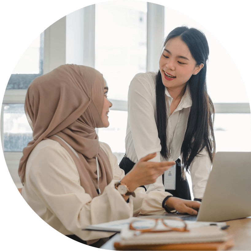 Two women in an office setting discuss work at a desk with a laptop. One wears a hijab, and both appear engaged in conversation.